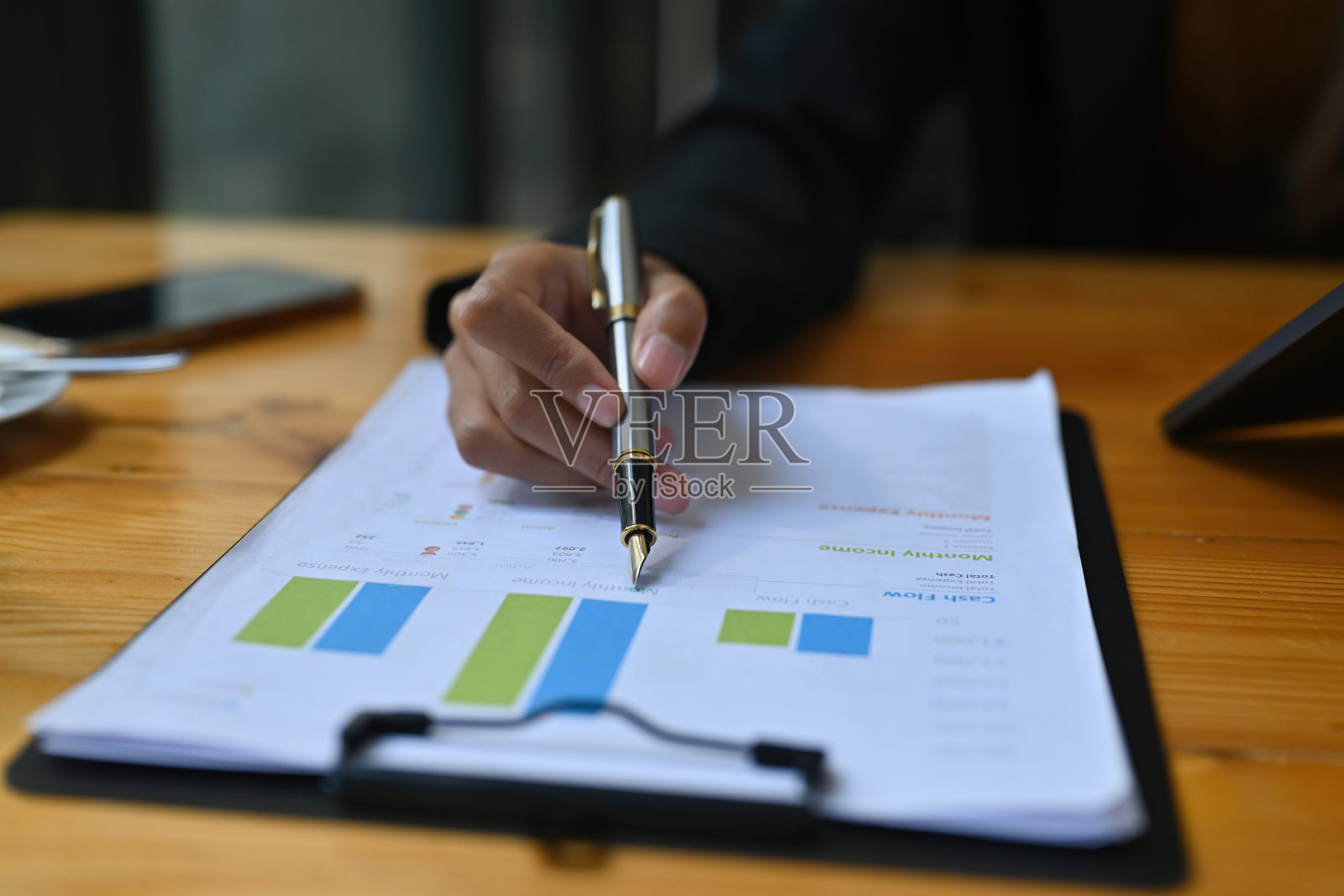 在被各种办公设备包围的木桌上，女商人手持一支笔，指着剪贴板检查业务信息的特写图像。照片摄影图片