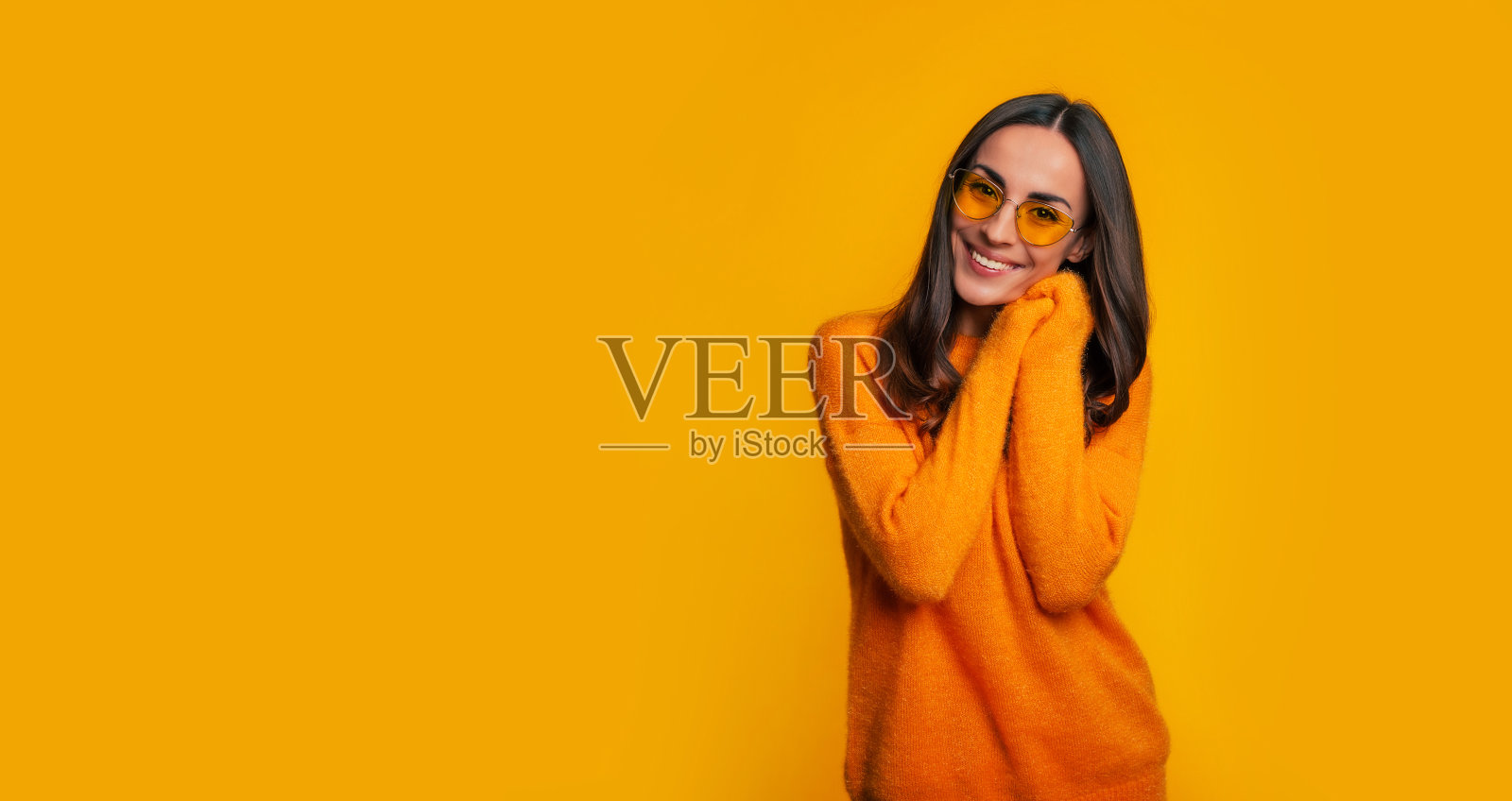 迷人的年轻女子在黄色毛衣和太阳镜，而她摆着微笑在黄色的背景下，看着相机照片摄影图片