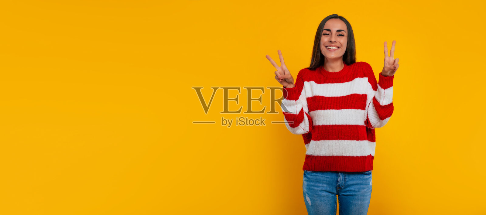 快乐微笑的美丽年轻的现代女性显示胜利的标志，而她摆孤立的黄色背景照片摄影图片