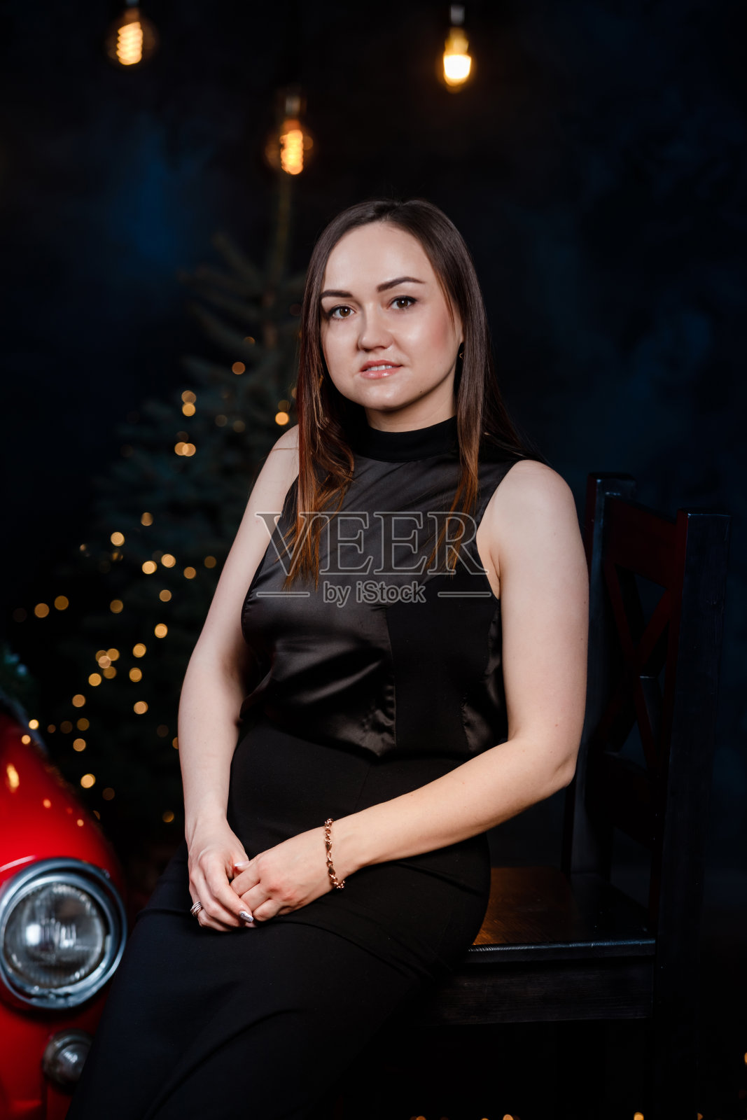 一个年轻美丽的女人穿着黑色的节日长裙站在圣诞树旁。优雅的女士在黑色连衣裙在圣诞树的灯光背景下。新年快乐。照片摄影图片