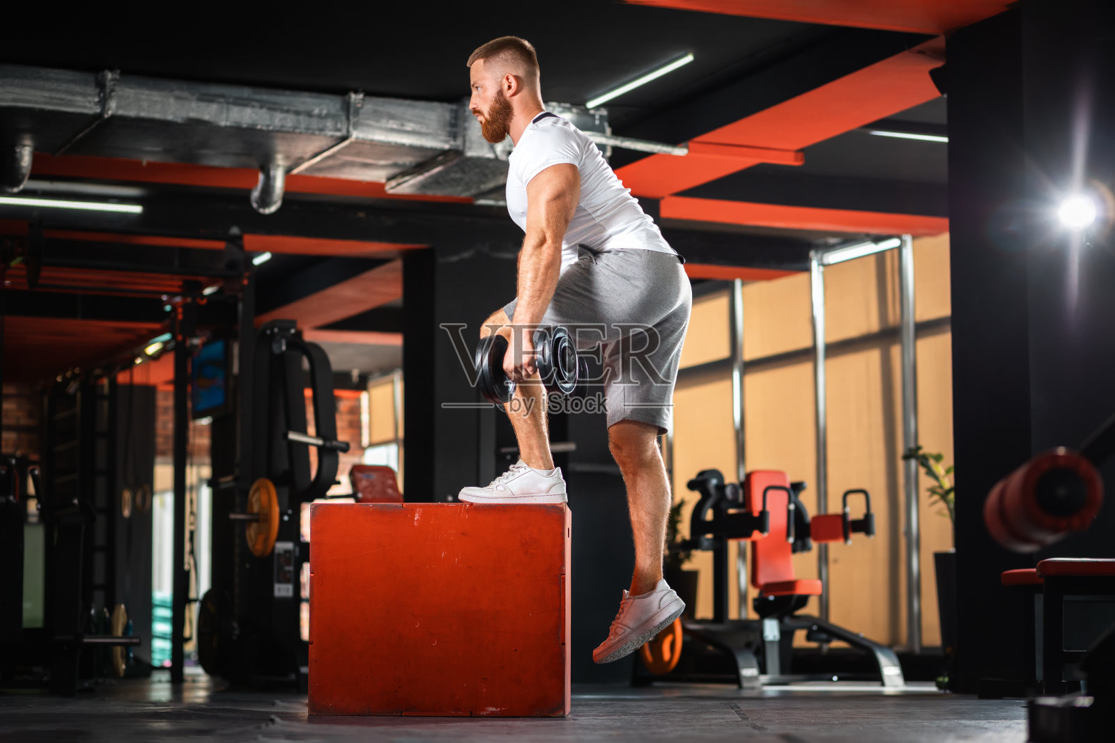 留着胡子的年轻运动员在健身房里训练拿着哑铃在立方体上跳跃。健身和健身的概念照片摄影图片