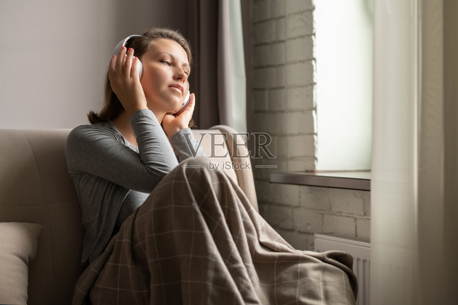 白人年轻女子的肖像，戴着耳机听音乐，坐在沙发上盖着毯子，从杯子里喝咖啡，看着窗户。照片摄影图片