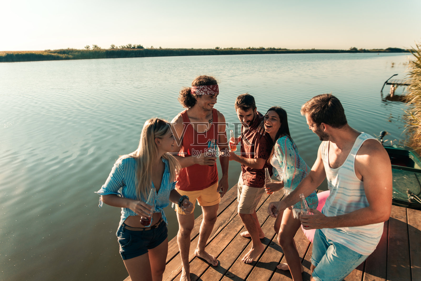 年轻的朋友们站在码头上，在湖边喝着酒精饮料干杯。照片摄影图片