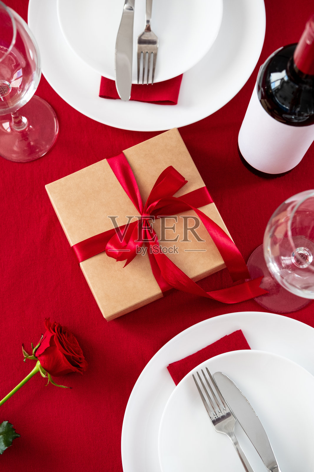 情人节的餐桌上摆放着餐具、礼品盒、酒瓶、玻璃杯、红玫瑰在红色的背景上。照片摄影图片