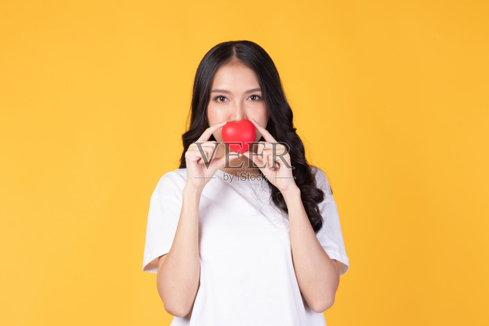 快乐亚洲年轻女子长卷发持有红色的心微笑可爱和可爱孤立在黄色的背景。爱和情人节。照片摄影图片