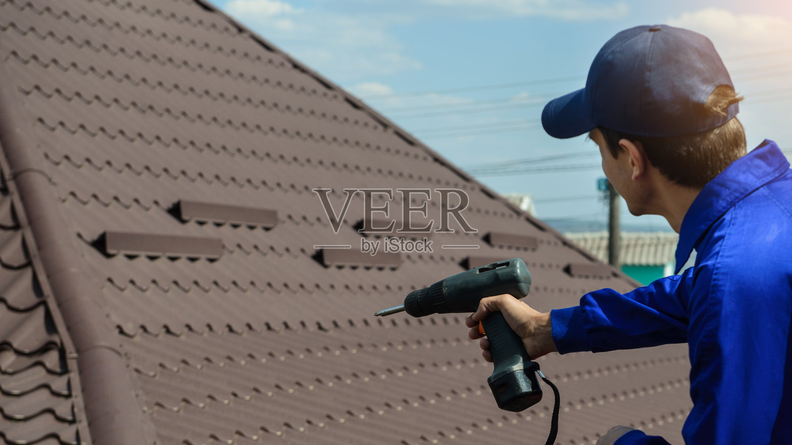 身穿蓝色工作服的承包商工人正在用电动螺丝刀修理棕色屋顶照片摄影图片