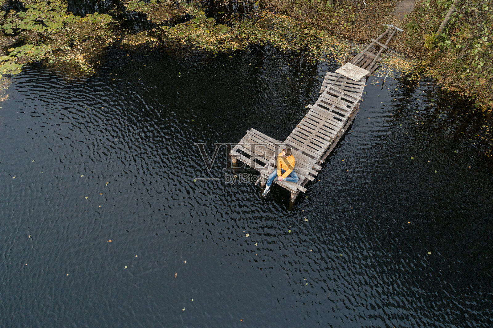 空中拍摄的一名女子，穿着黄色夹克，放松地躺在湖边的木制码头上，独自观看秋天的日落。照片摄影图片