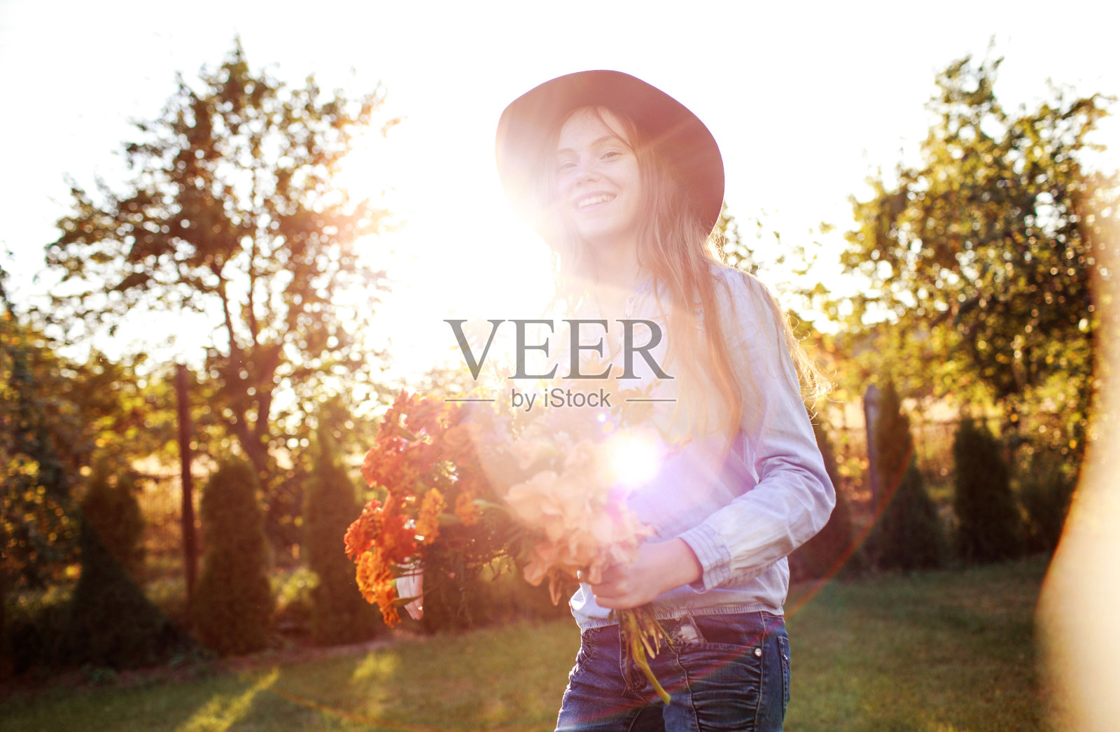 在夕阳的映衬下，一个头戴红帽子的红发少女手捧一束鲜花在草坪上翩翩起舞照片摄影图片