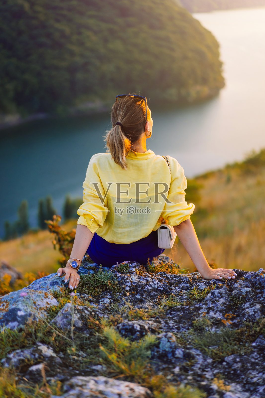 一个年轻时髦的女人坐在悬崖边的岩石上。徒步旅行。神奇的自然，夏日和日落。认不出来旅行的女孩。冒险的生活方式照片摄影图片