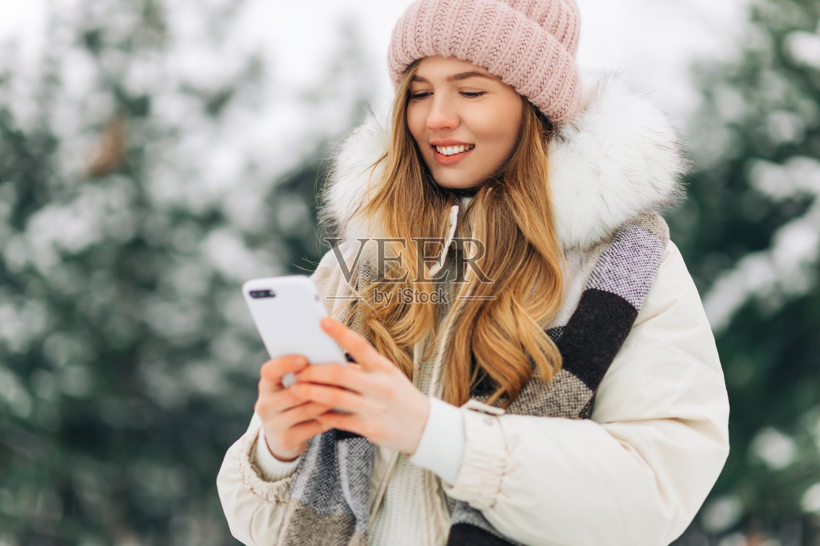 女人戴着针织帽，穿着暖和的外套，在冬季公园散步，用手机照片摄影图片