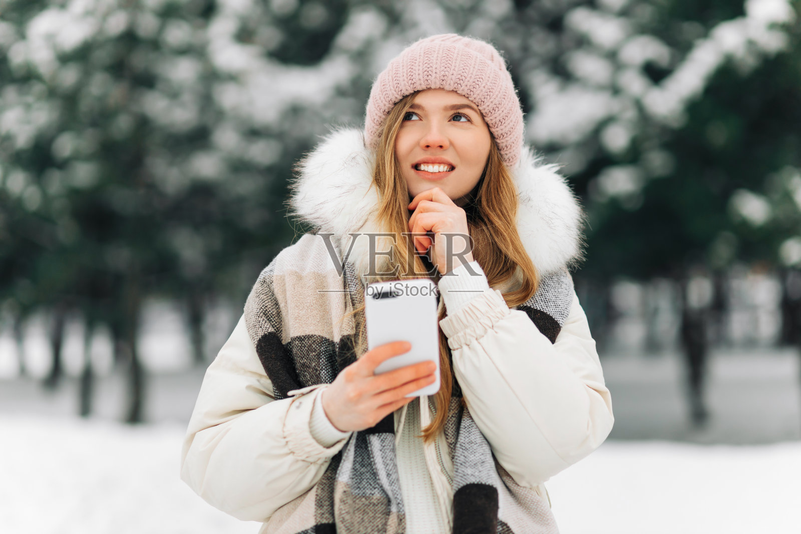 冬天，戴着暖和的针织帽和波尔托毡帽的沉思的女人在户外使用智能手机照片摄影图片