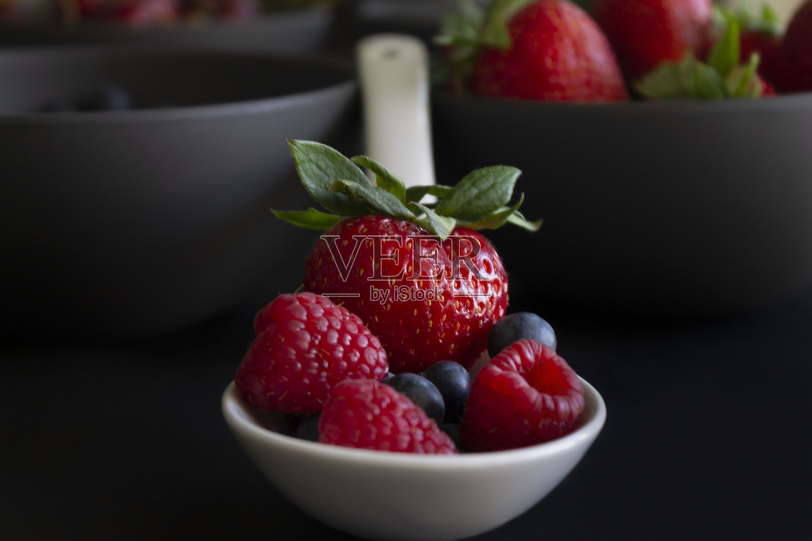 浆果装在白勺和四个碗里的创意食物概念。新鲜草莓、蓝莓和覆盆子。新鲜的森林水果在黑色的背景。照片摄影图片