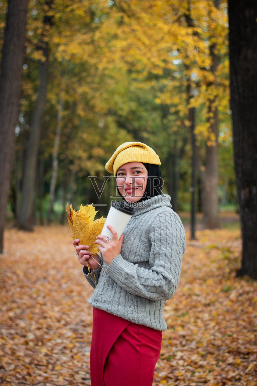 年轻的穆斯林妇女戴着黄色的帽子，穿着灰色的毛衣，一杯咖啡和一片黄色的秋叶。秋天的公园。女人味和微笑。照片摄影图片