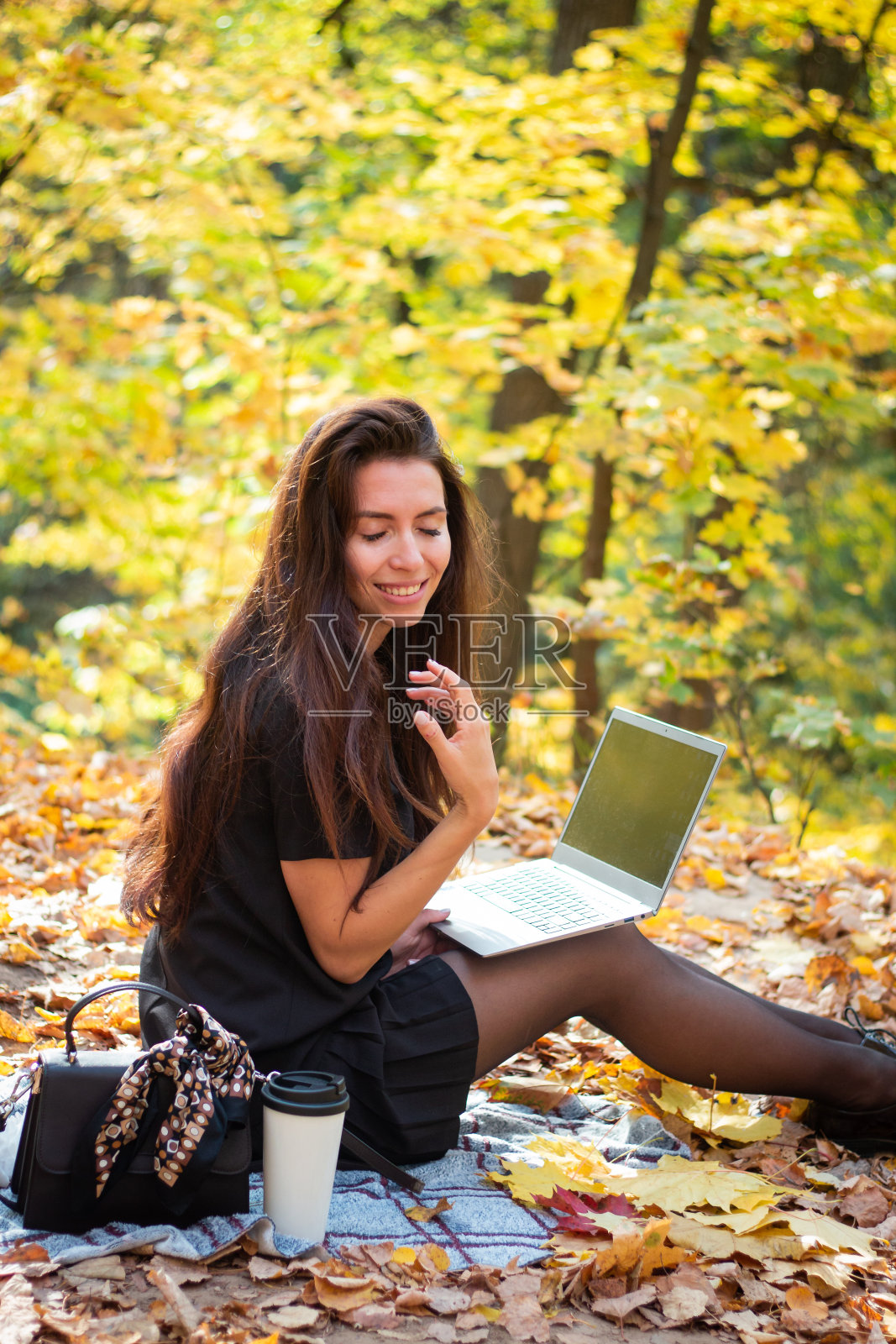 一个深色头发的女人正坐在秋天的公园里，手里拿着一台笔记本电脑和一杯咖啡。学生。老师。在线教育照片摄影图片