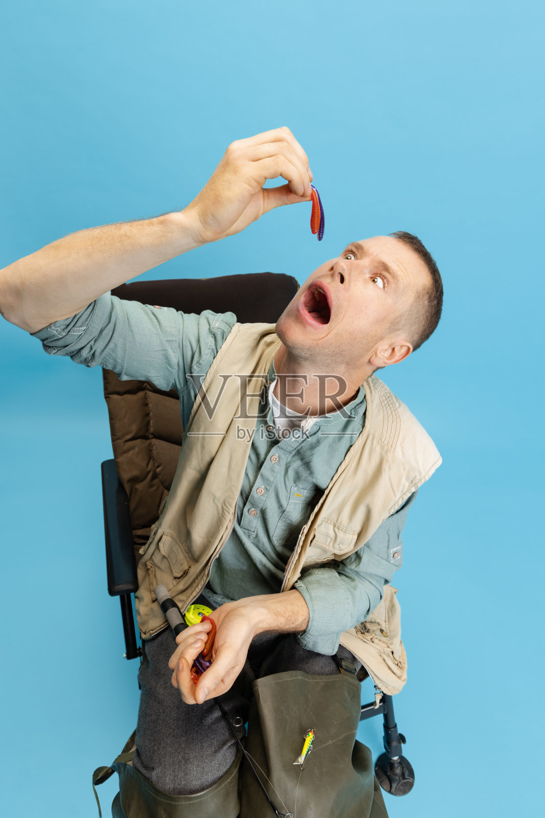 有趣的年轻人，渔民与钓鱼配件坐在椅子上孤立的蓝色工作室背景照片摄影图片