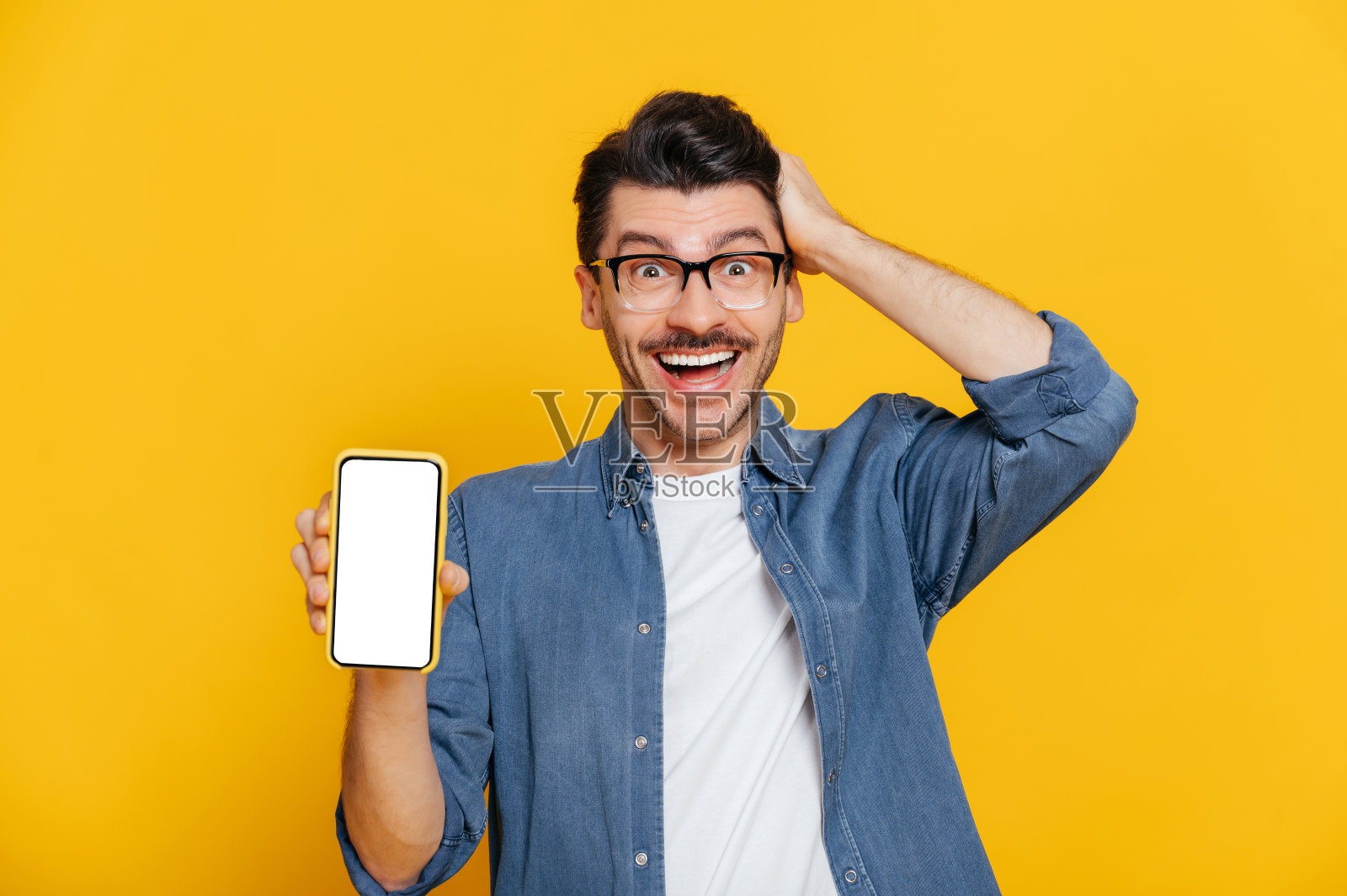 一名白人男子穿着牛仔衬衫，戴着眼镜，手持智能手机，用手触碰头部，站在孤立的橙色背景下，看着相机照片摄影图片