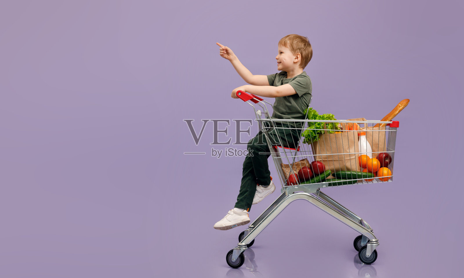 一个男孩坐在购物车里指着他的手指。与cjpe空间购物的孤立概念图像。照片摄影图片