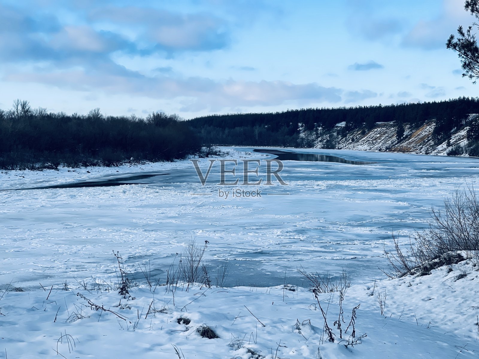 森林之间白雪覆盖的河流的冬季景观。在冬季多云的天气里，结冰的河流的美丽景色照片摄影图片