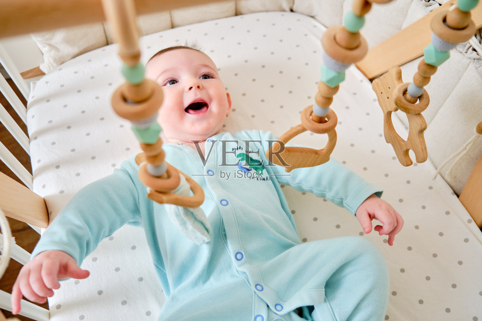 快乐的婴儿宝宝正躺在床上玩着木制的挂玩具。一个六个月大、穿着绿松石色衣服、面带微笑的滑稽孩子躺在婴儿床上照片摄影图片