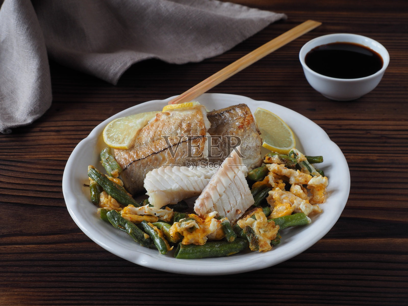 煎黑线鳕片，白色盘子里的青豆和鸡蛋，酱油，深色木桌上的筷子，平面布置。饮食菜以蔬菜和海鲜为主照片摄影图片