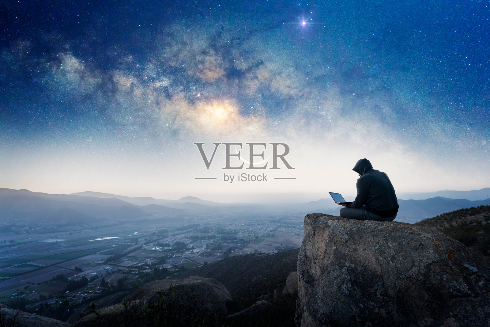 一个人坐在山顶上的剪影，背景是一台笔记本电脑和银河照片摄影图片