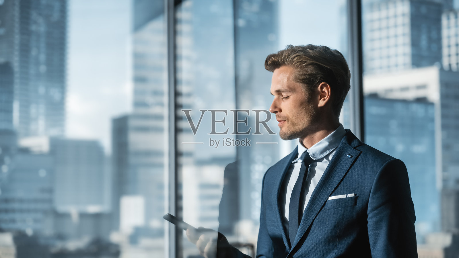 穿着西装的自信的年轻商人站在现代的办公室里，用智能手机，看着窗外有摩天大楼的大城市。成功的财务经理计划工作项目。照片摄影图片