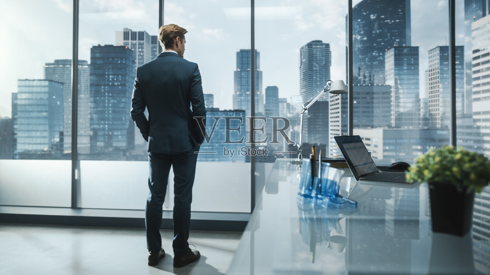 年轻成功的商人，穿着考究的西装，站在他的现代办公室里，看着窗外高楼林立的大城市。成功的财务经理计划项目策略。照片摄影图片