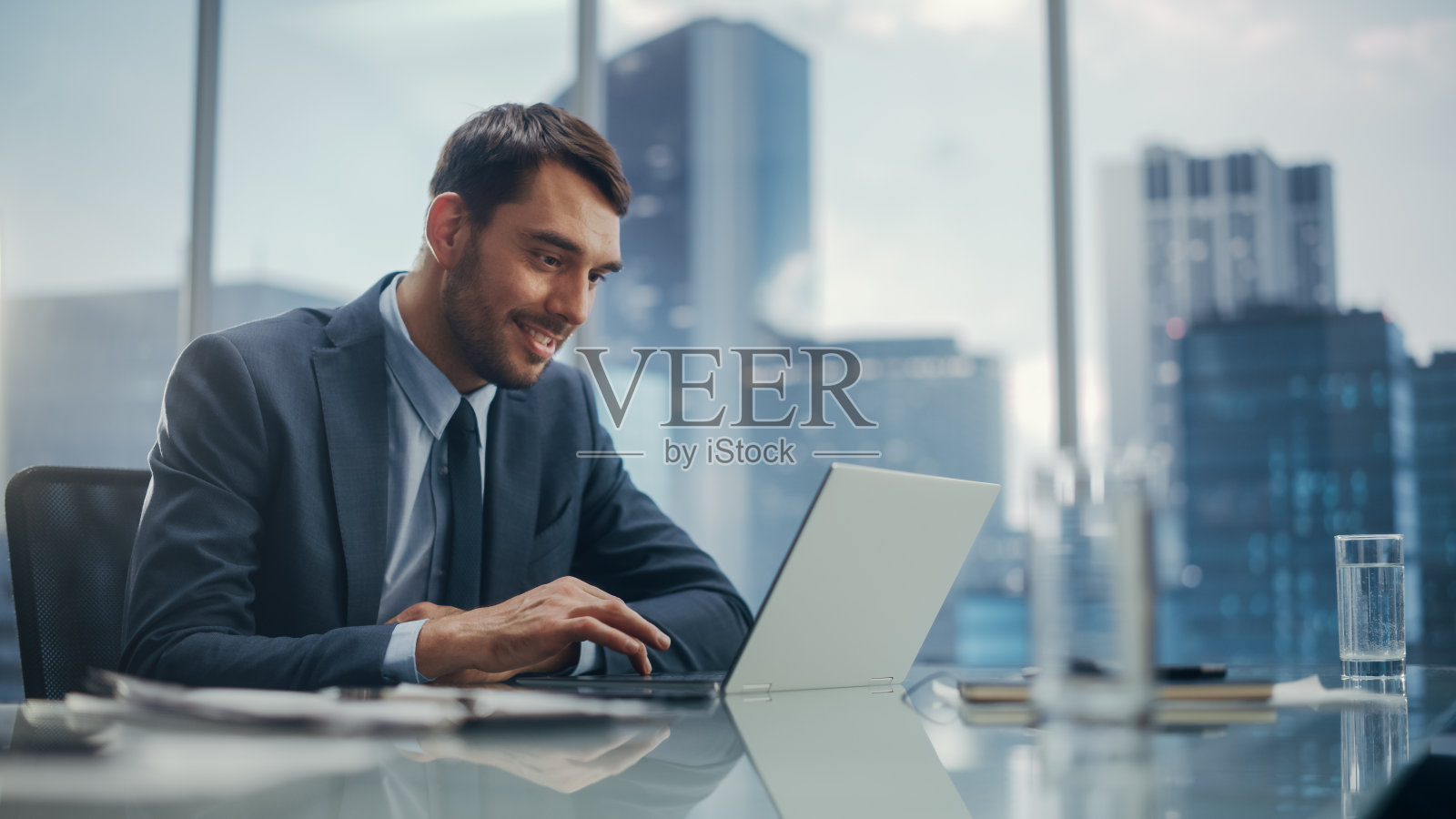 快乐的西装商人坐在现代办公室的办公桌前，使用笔记本电脑，紧靠窗外的大城市和摩天大楼。成功的财务经理计划工作项目。照片摄影图片