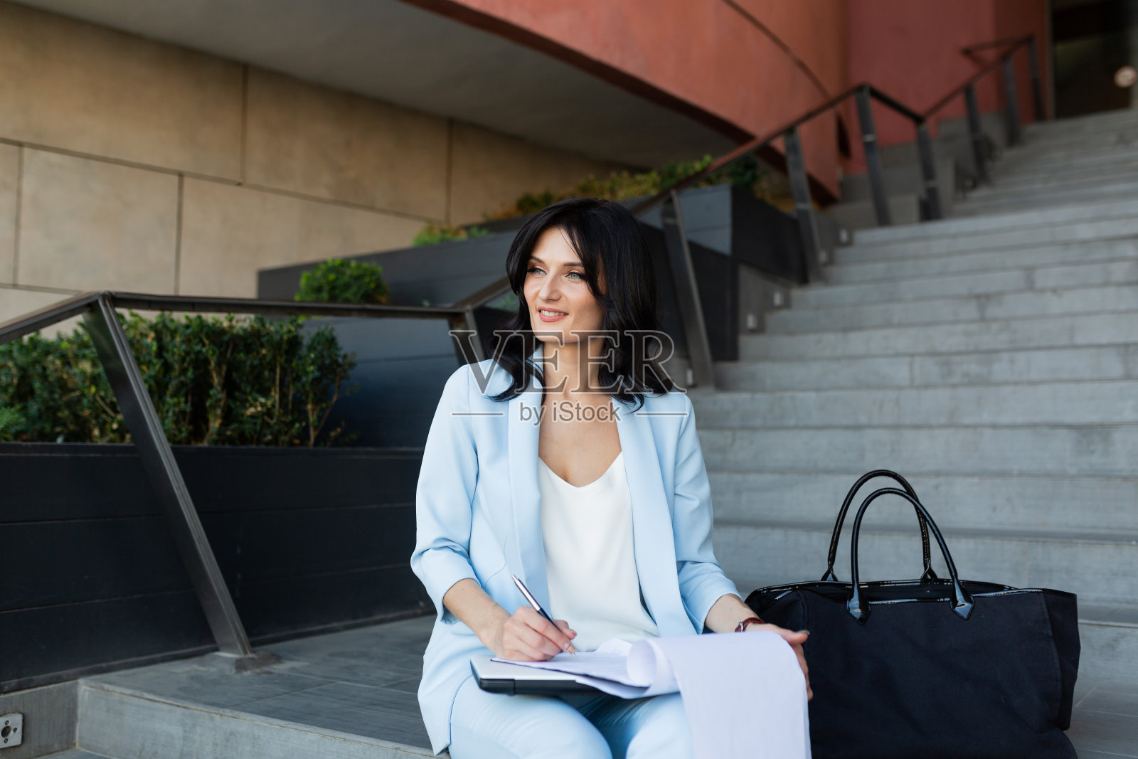 年轻漂亮的女商人在办公中心附近的楼梯上用笔记本电脑和文件工作，打电话照片摄影图片