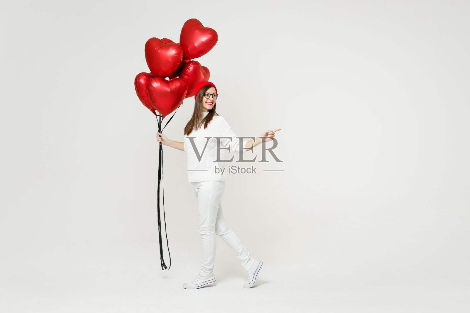 全长度侧面的有趣的年轻女子在毛衣红色帽子眼镜点手指一边庆祝生日节日派对持有一束心充气气球孤立在白色背景。照片摄影图片