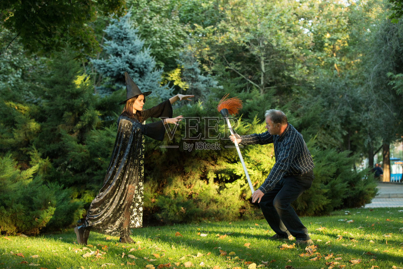 一个年轻漂亮的女孩穿着女巫的服装，正试图迷惑一个手里拿着花园扫帚的男人。万圣节。照片摄影图片