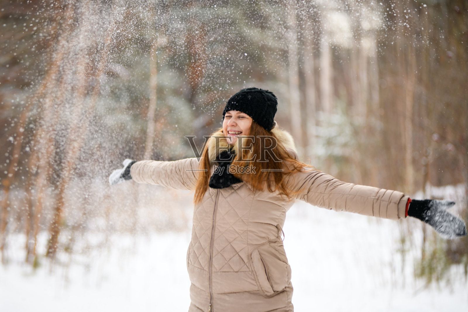 红发女孩把雪扔到顶部，微笑在冬季的森林里，穿着暖和的衣服:黑色的帽子，围巾和手套和米色的夹克照片摄影图片