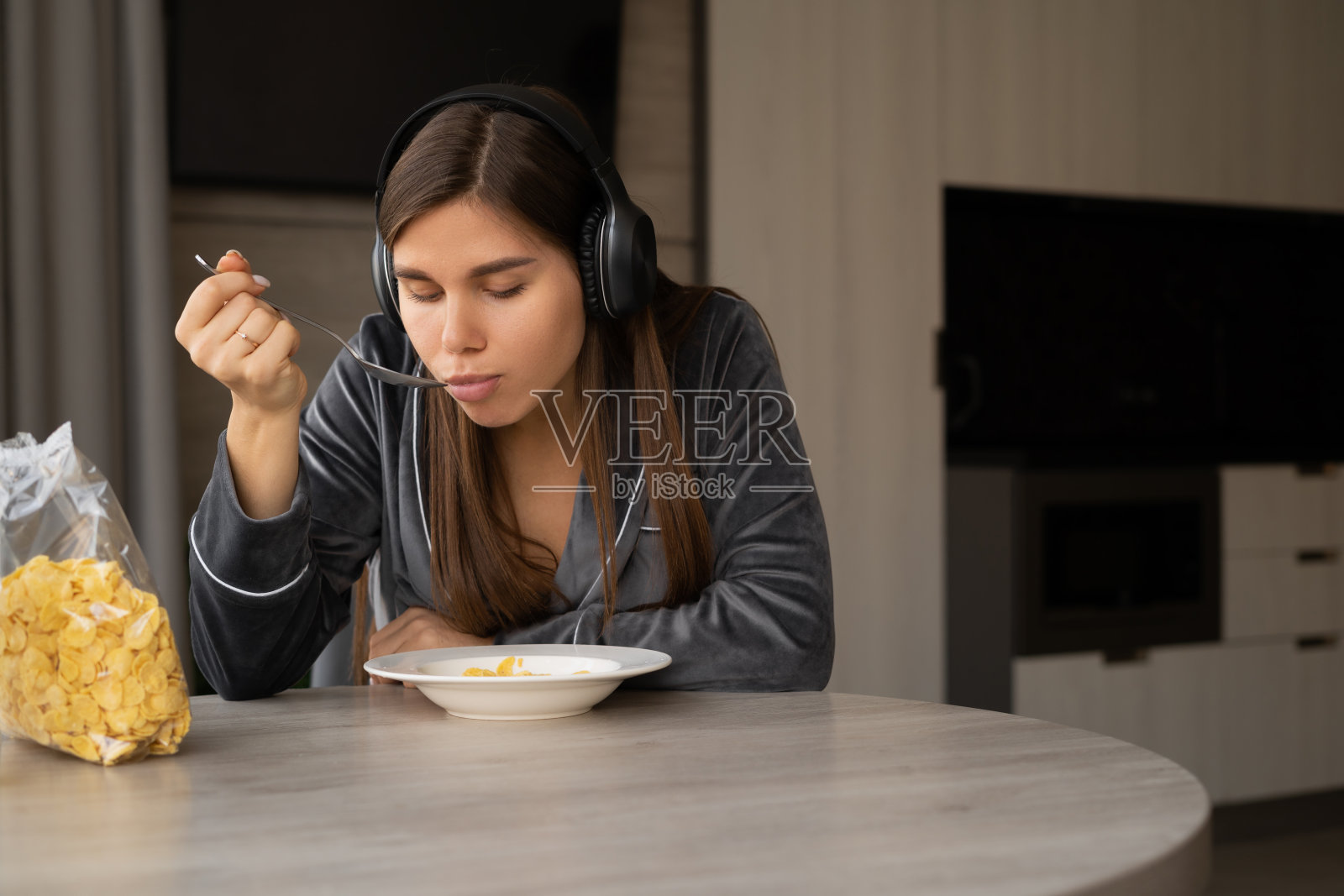 坐在家里拿着碗和勺子吃麦片的快乐女人。早餐时戴上无线耳机听音乐。照片摄影图片