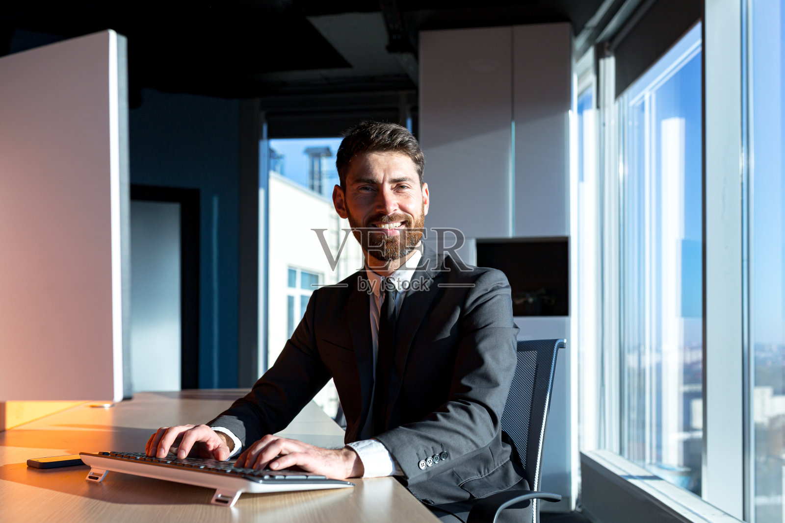快乐、微笑的商人在现代办公室里靠窗工作，大胡子老板照片摄影图片