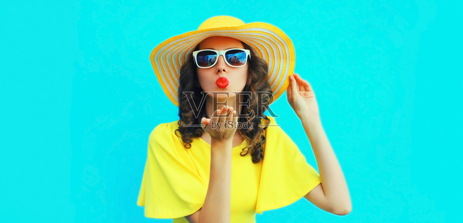 美丽的年轻女子吹着她的嘴唇送甜蜜的空中吻的肖像，戴着黄色夏天草帽，穿着蓝色的背景照片摄影图片