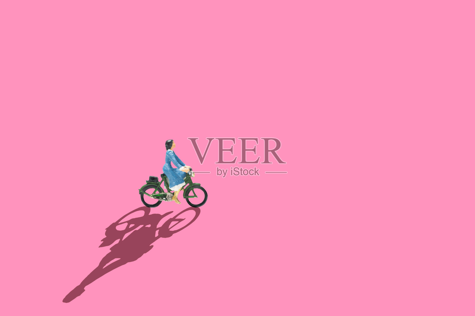 选择性聚焦微型女性蓝色连衣裙骑自行车在粉红色的背景和长阴影。照片摄影图片