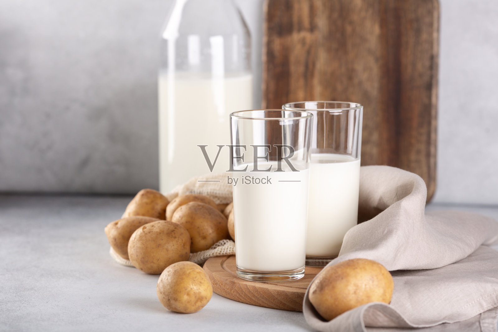 纯素植物牛奶装在两个透明的玻璃杯和瓶子里。灰色的桌子上放着土豆奶和土豆块茎照片摄影图片