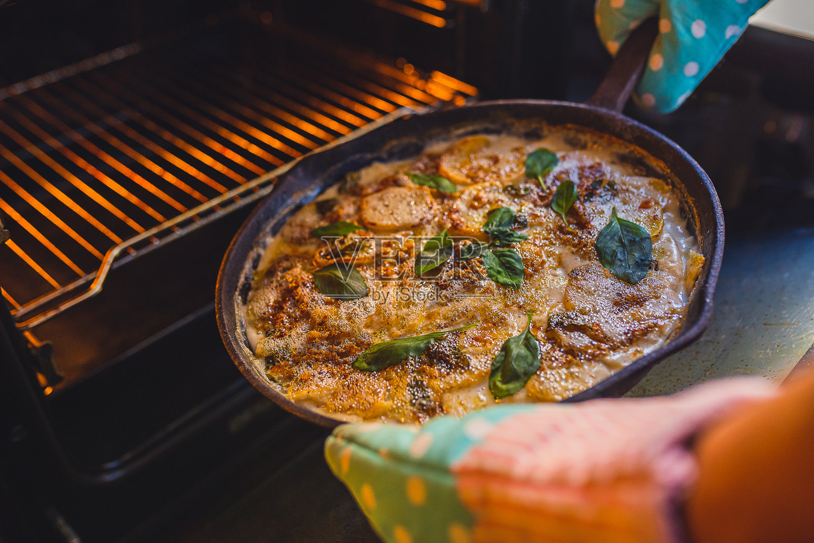 装在铸铁煎锅里的两次烤土豆，上面放着磨碎的奶酪、罗勒和培根，双手戴着厨房手套照片摄影图片