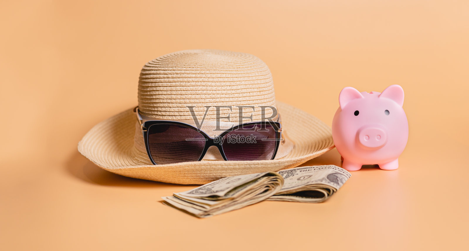 为旅游省钱的理念。一个女太阳帽，太阳镜，美元钞票和一个储钱罐在橙色的背景上。照片摄影图片