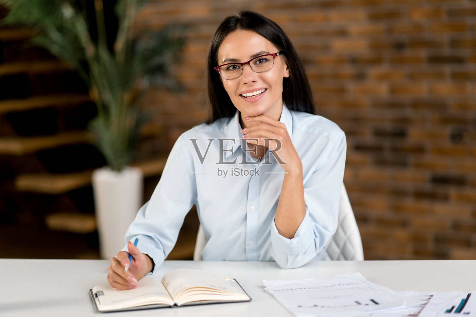 美丽的聪明的成功的年轻成年黑发白人女商人，首席执行官或经纪人戴眼镜，坐在现代办公室的办公桌，看着相机，微笑友好照片摄影图片