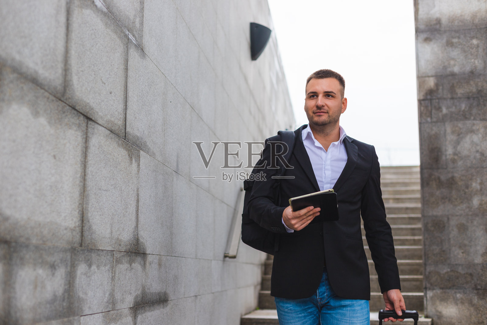 一个年轻的白人商人背着双肩包，拿着一台数码平板电脑和一个手提箱走下楼梯照片摄影图片