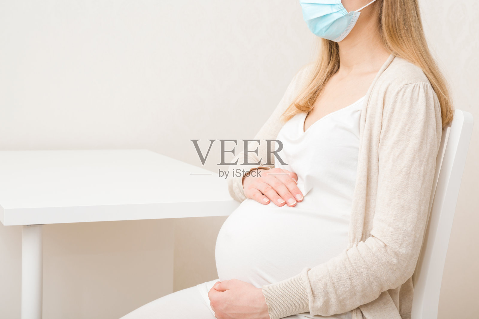 年轻成年孕妇戴着口罩，坐在妇科医生办公室的桌子旁。怀孕期间定期检查。双手触碰大肚子。侧视图。特写镜头。照片摄影图片