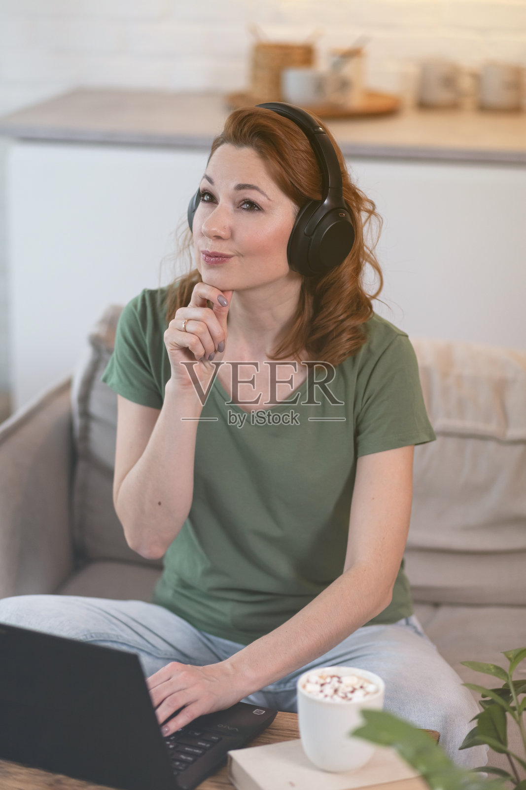 美丽的微笑红发年轻女子远程工作在家里厨房在她的笔记本电脑，听音乐。远程自由职业的概念照片摄影图片