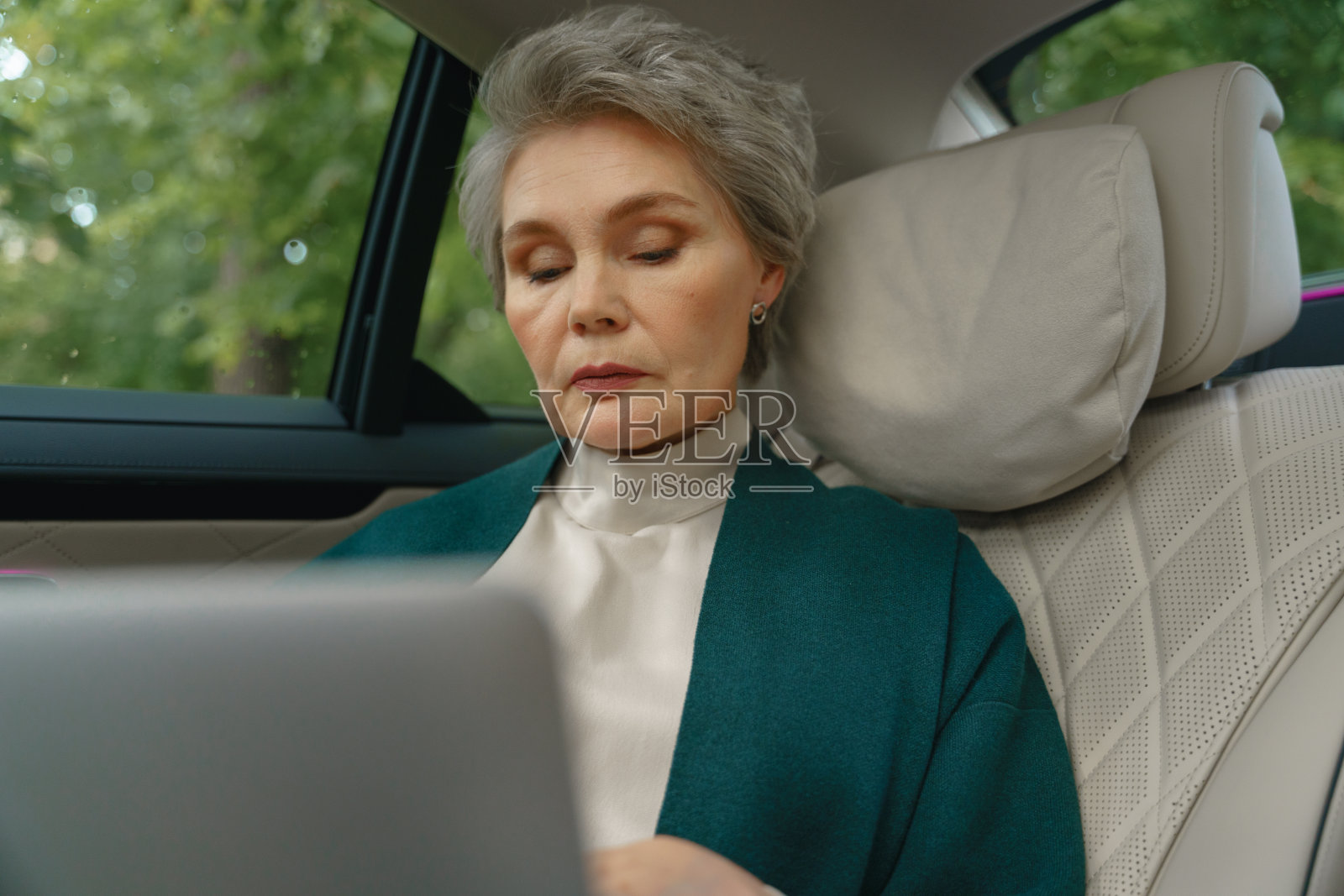 一位漂亮的高级商务女性，头发花白，化着专业的妆，坐在一辆豪华轿车的副驾驶座位上，在大腿上用笔记本电脑工作照片摄影图片