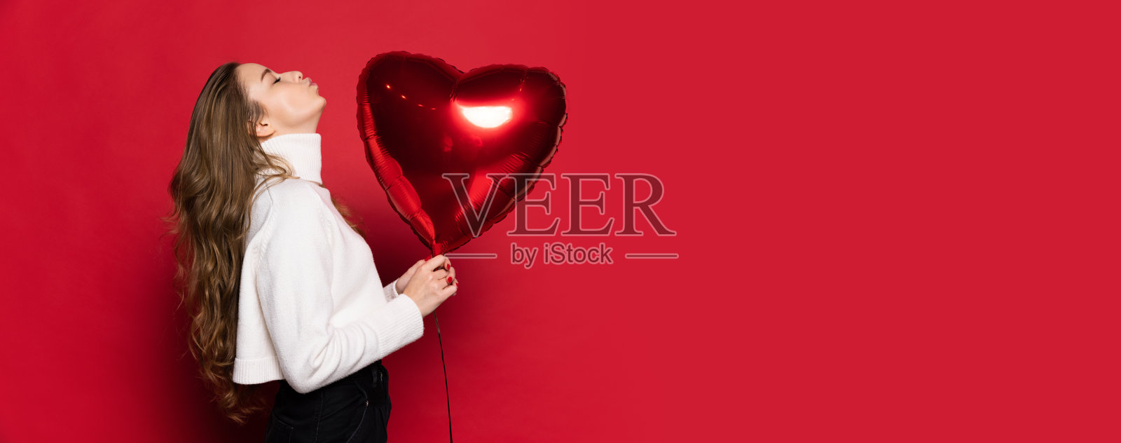 年轻漂亮，长波浪丝般的头发，自然的化妆拿着红色的心气球仰望孤立在红色的背景。情人节的概念照片摄影图片