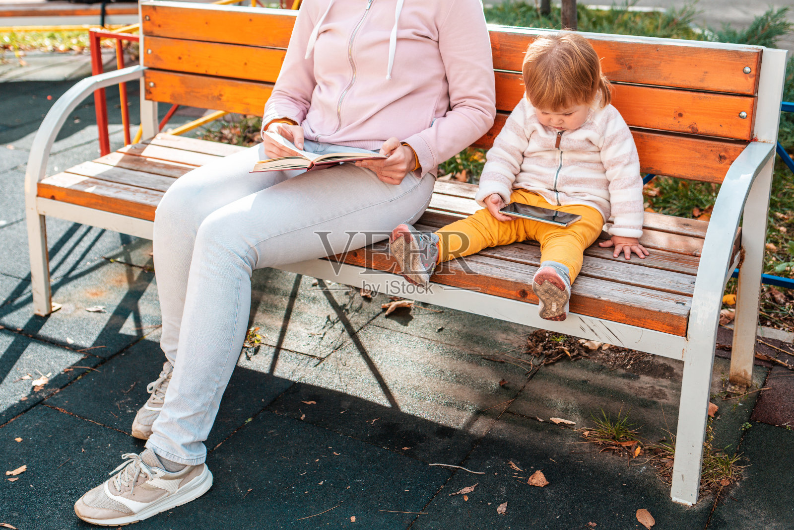 母亲和孩子坐在长椅上，一边看书一边玩手机放松身心。公园里温馨的家庭休闲时光照片摄影图片