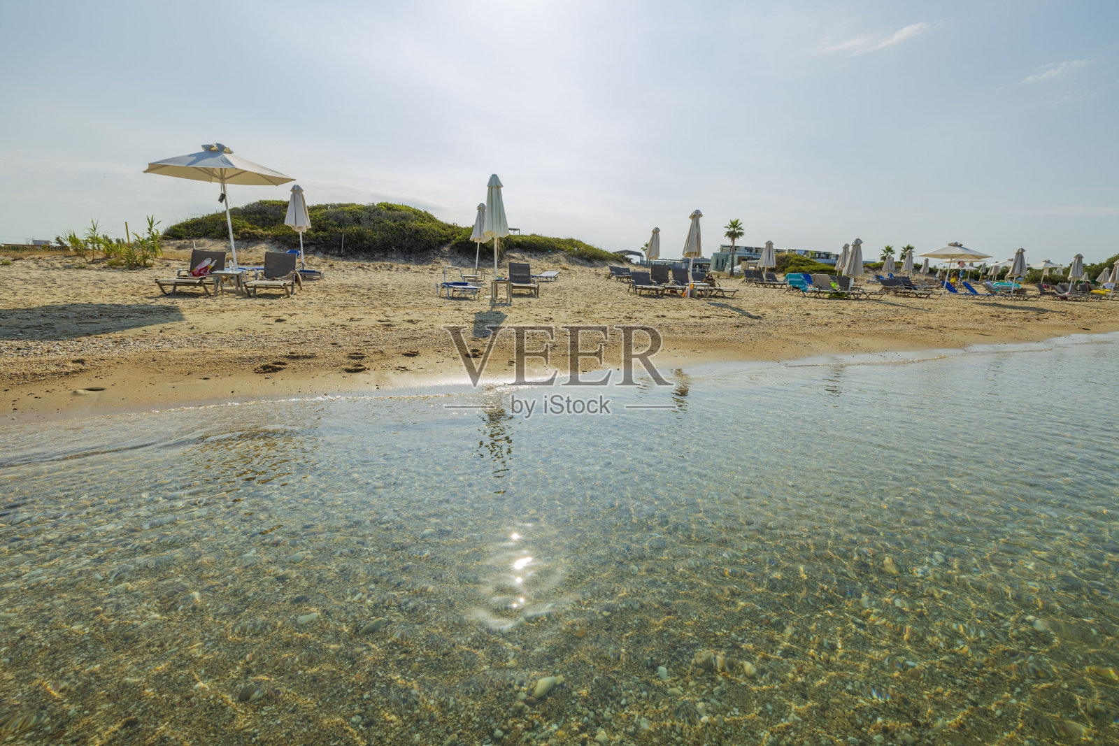 美丽的白色沙滩希腊海滩，阳光伞下的空日光浴床。镜面般清澈的海水表面上的太阳反射。照片摄影图片