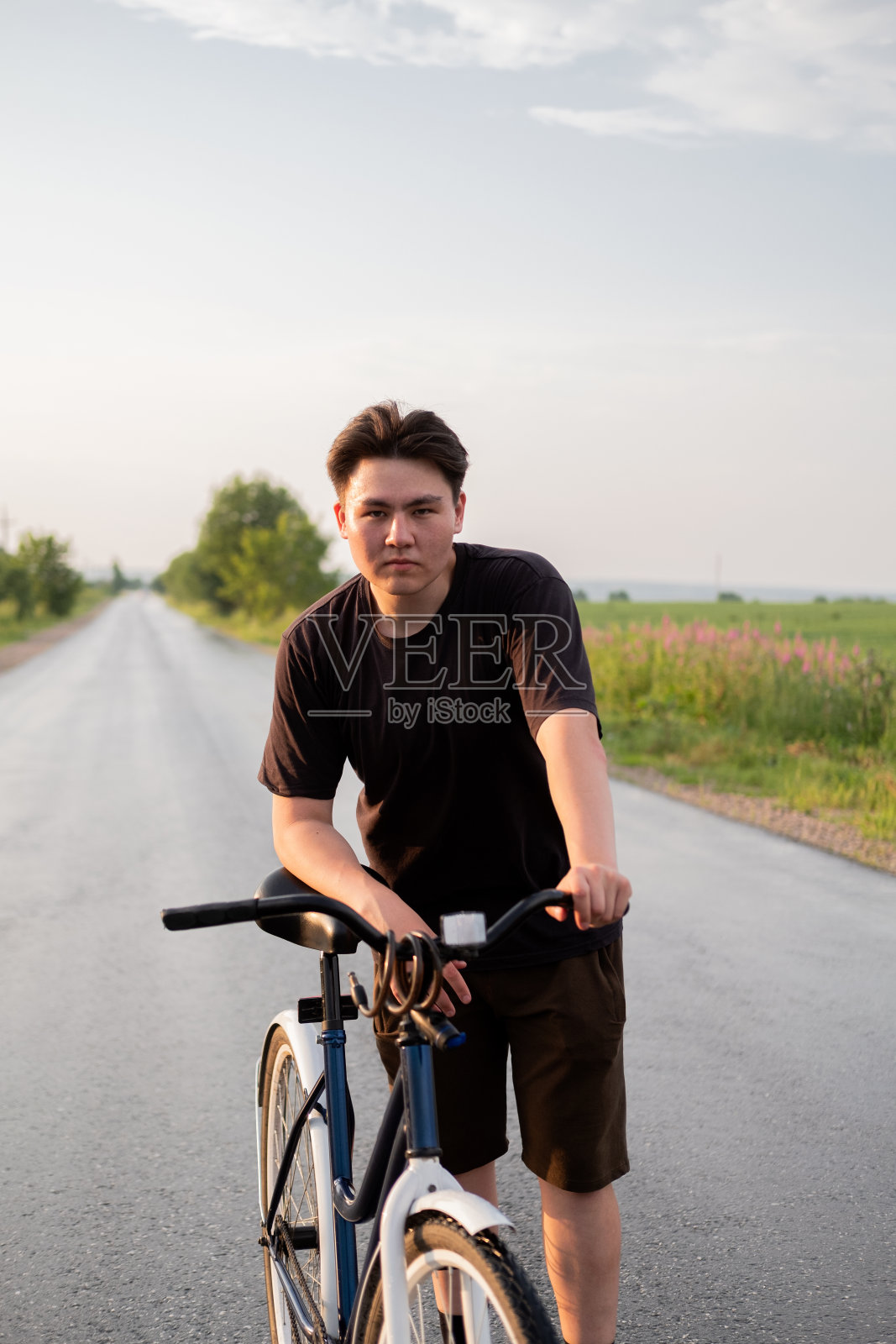 年轻人独自站在路上，骑着自行车，看着镜头，看着乡村。夏季活动，健康的生活方式，锻炼，运动，健身。人在自然界中。骑自行车。在户外玩。照片摄影图片