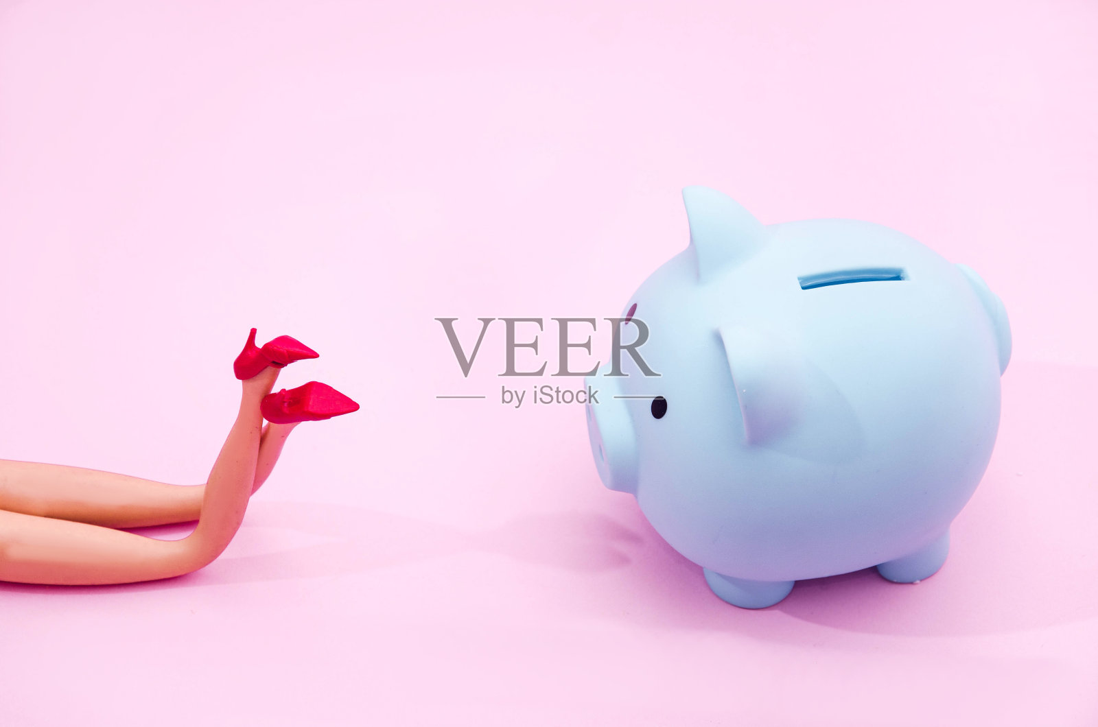 穿着红色鞋子的女性的腿和一个蓝色小猪存钱罐，粉色粉彩背景。情人节banner或卡片的创意概念。爱与金钱商业概念的艺术设计照片摄影图片