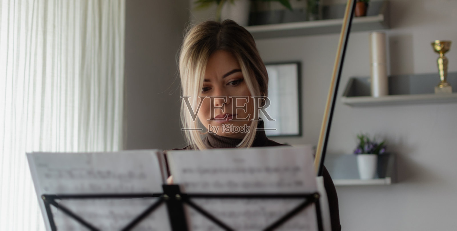 美丽和年轻的女孩享受在她的家里演奏她的中提琴，练习和享受她的自由时间，在阳光明媚的日子里在窗户附近演奏。照片摄影图片